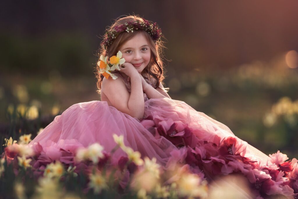 Young girl in a flower crown and pink princess gown sitting in a field of daffodils at golden hour
