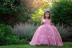 irl in sparkling pink gown and crown holding a magic wand in a garden setting.