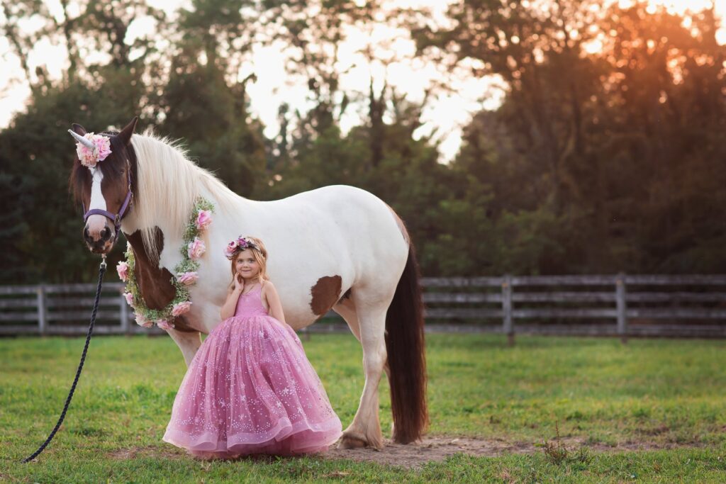 Girl in pink dress standing beside a flower-adorned white horse at sunset.