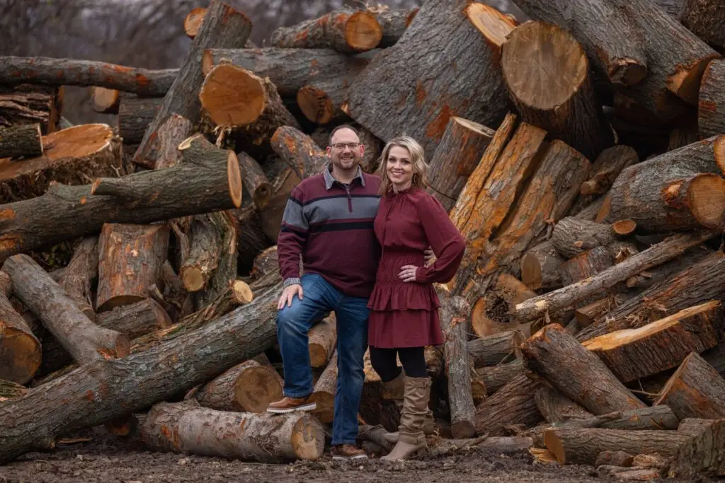 Smiling couple poses in front of a pile of large cut wood, wearing burgundy and earth-tone outfits—ideal for a rustic-themed holiday photo session.