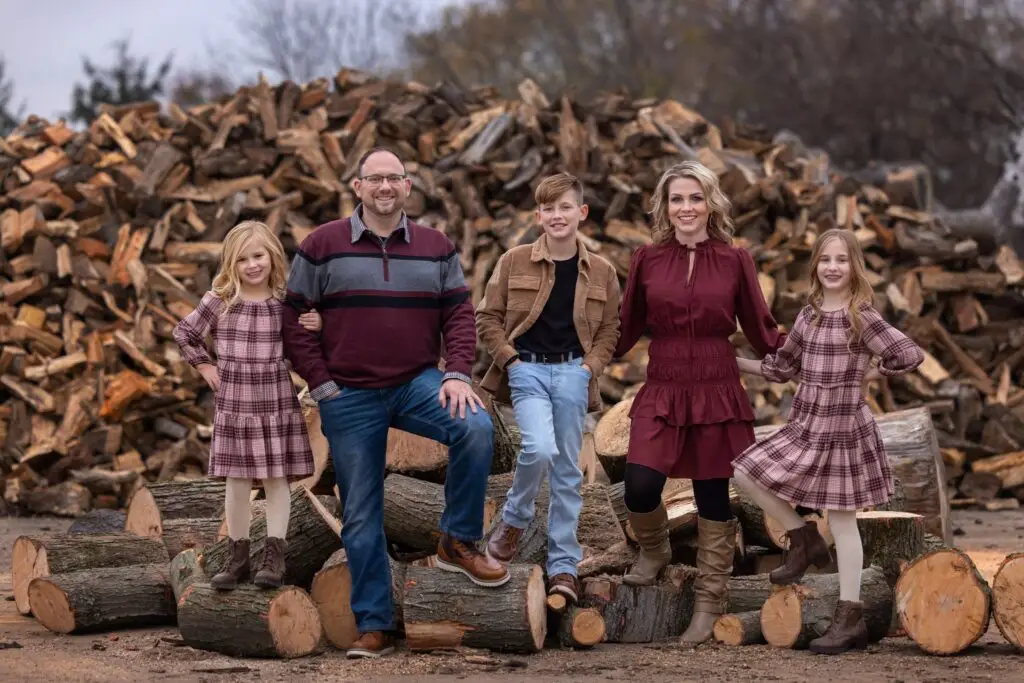 Family of five smiles in front of a woodpile, dressed in matching burgundy, flannel, and neutral tones—great inspiration for outdoor holiday family photos.