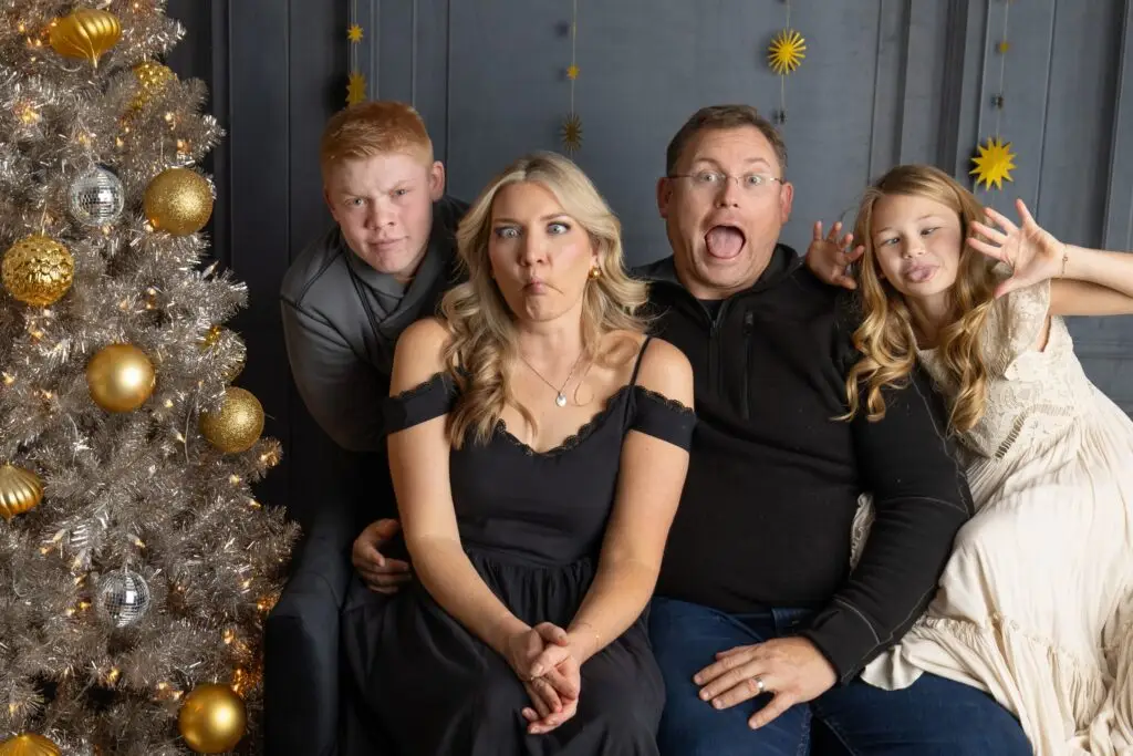 Fun Christmas family photo with teens in studio holiday setup – Cream N Cocoa Family of four poses with silly faces during a festive holiday photo session in a studio decorated with gold ornaments and a silver Christmas tree, great for capturing teen personalities.