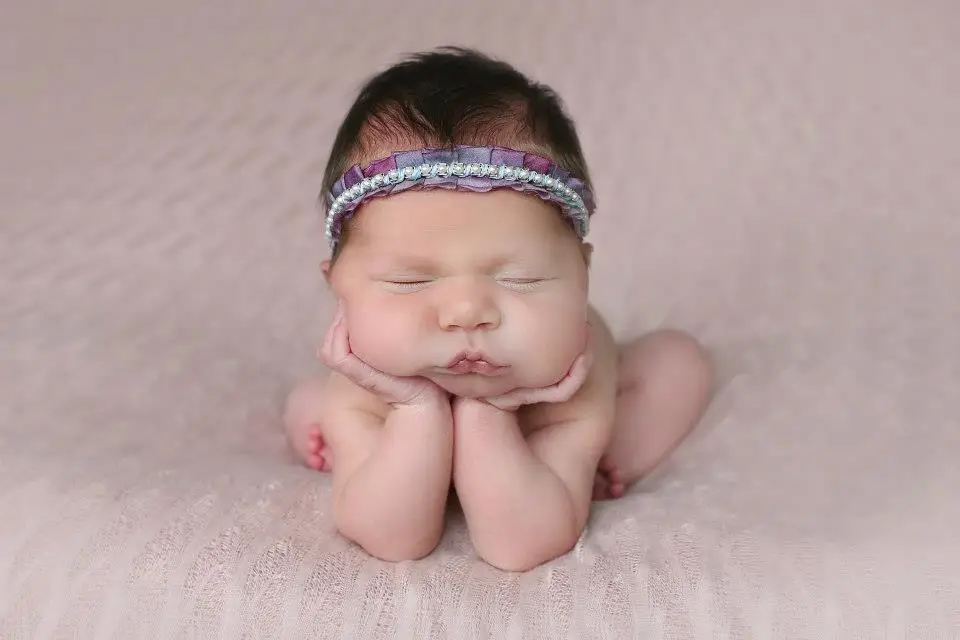 Newborn baby girl posed with hands under chin on soft pink fabric, wearing a purple headband