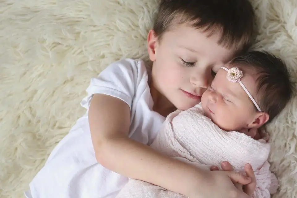 Older brother holding newborn sister while lying on a cream fur blanket, both with closed eyes