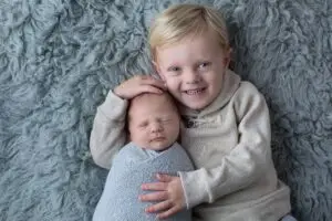 Older brother smiling while gently holding newborn baby swaddled in blue on fluffy gray blanket during a Cincinnati newborn photography session.