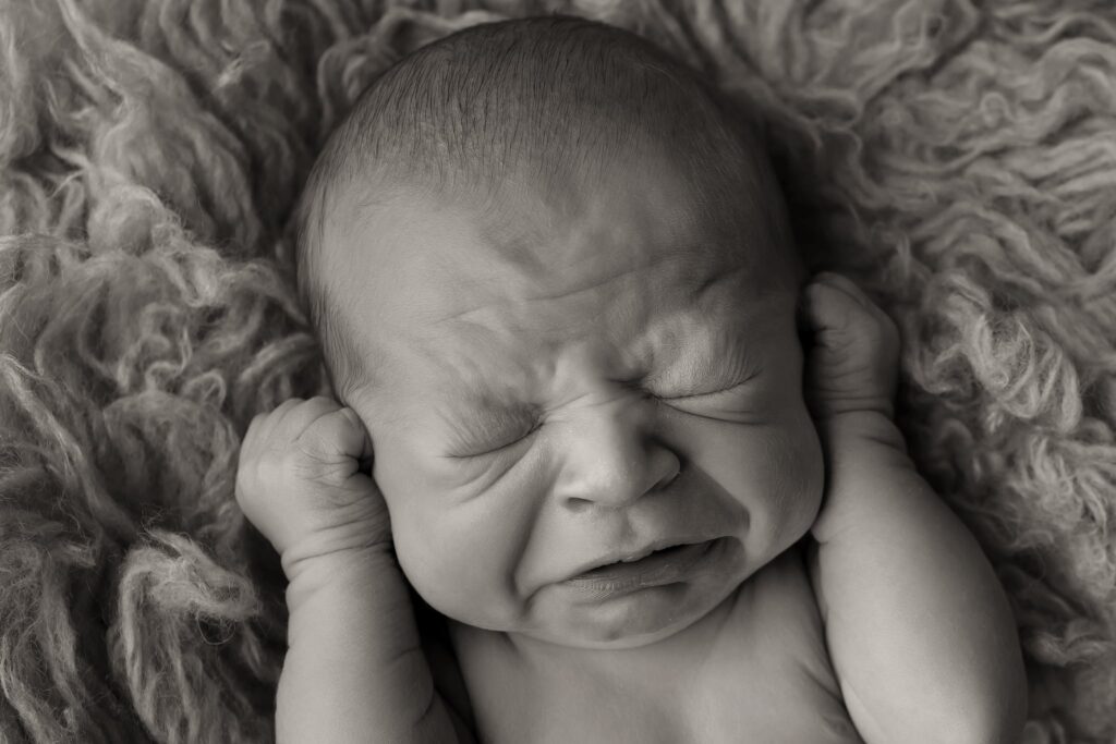 Black and white close-up of a newborn mid-sneeze with hands near ears, captured during a candid moment in a Cincinnati newborn photography session.