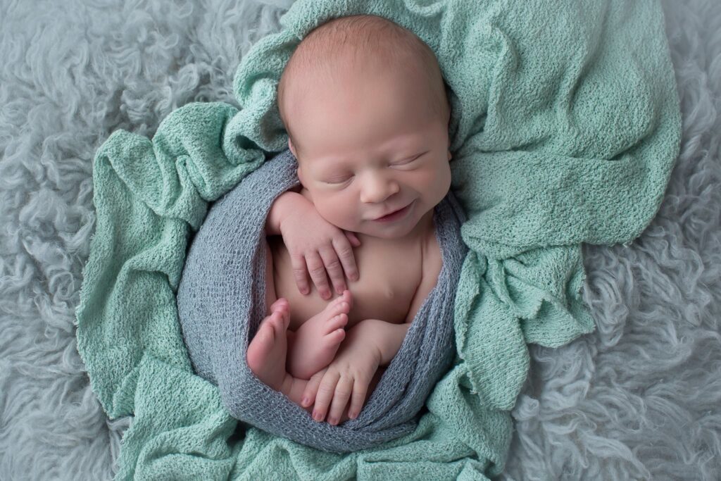 Newborn baby wrapped in gray and mint green blankets, softly smiling with hands and toes visible, photographed in a Cincinnati newborn studio.