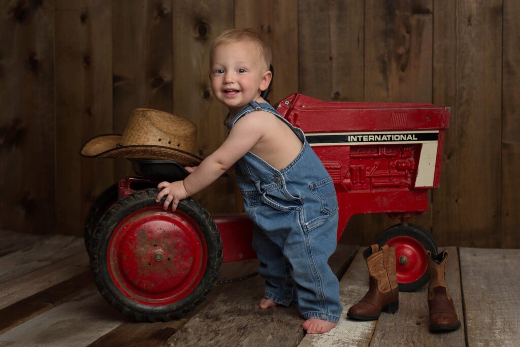 One-year-old boy standing barefoot in denim overalls beside a red vintage tractor, with a cowboy hat and boots nearby during his first birthday photo session.