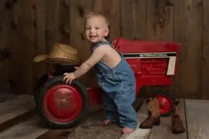 One-year-old boy standing barefoot in denim overalls beside a red vintage tractor, with a cowboy hat and boots nearby during his first birthday photo session.