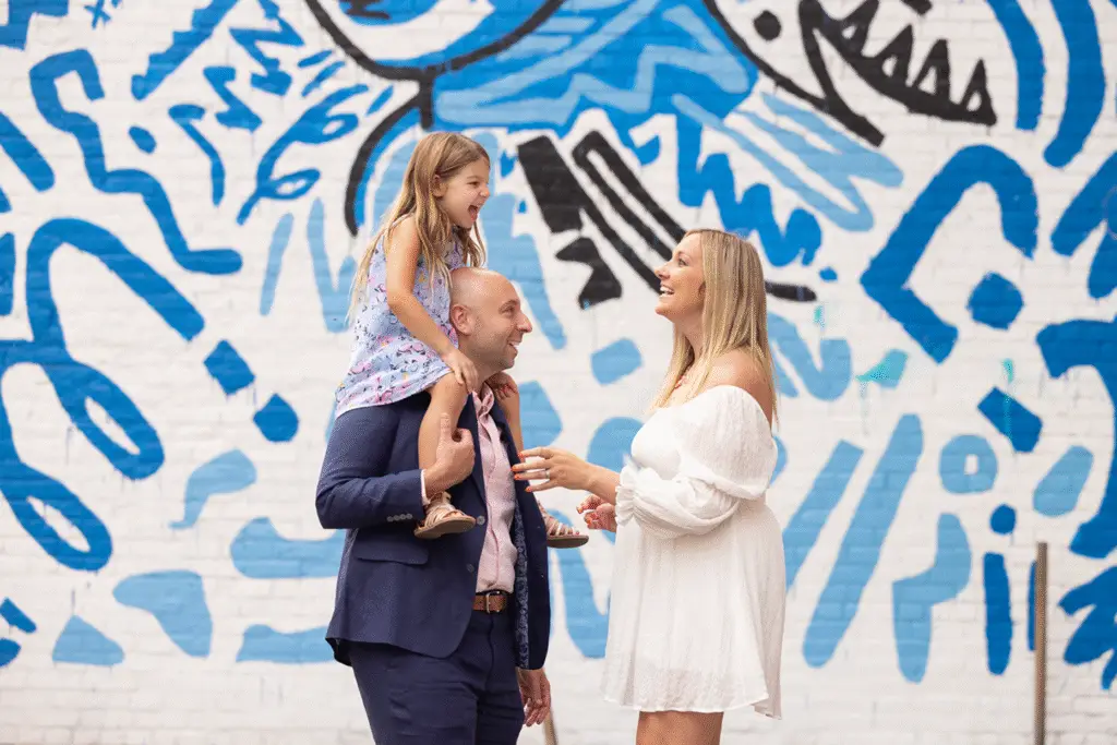 Smiling family during an urban outdoor photo session in Cincinnati, Ohio, featuring a father holding his daughter on his shoulders as they laugh with the mother in front of a colorful blue mural.