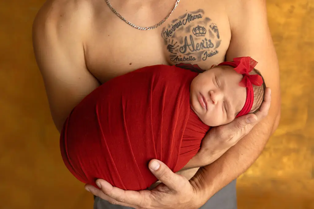Newborn photographer in Cincinnati capturing a sleepy baby wrapped in red, cradled safely in father’s arms