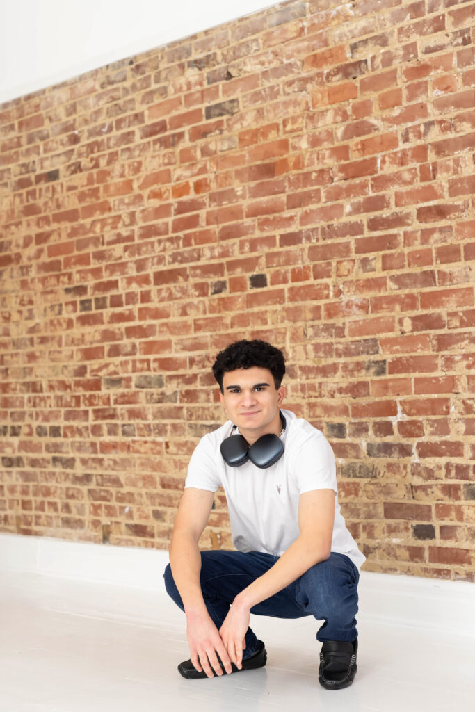 Young man crouching in front of a brick wall, wearing a white tee and headphones around his neck for his Senior portraits in Cincinnati