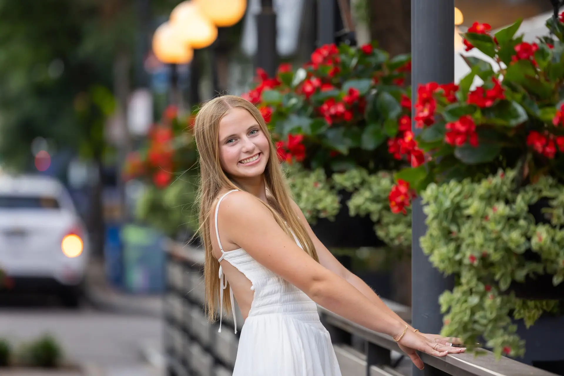 Smiling woman in a white dress leaning on a railing beside a wall of red flowers on a city street. posing for her Senior portraits in Cincinnati