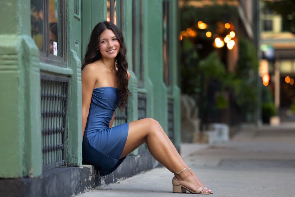 Smiling teen in a blue off-the-shoulder dress sits leaning against a green storefront on a city sidewalk, beige sandals visible posing for her Senior portraits in Cincinnati